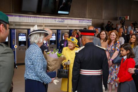 Her Majesty the Queen dressed in yellow coat 和 hat, with a school girls in red sweater, 一个穿蓝色/红色制服的人, a lady in blue with hat 和 other members of the public.